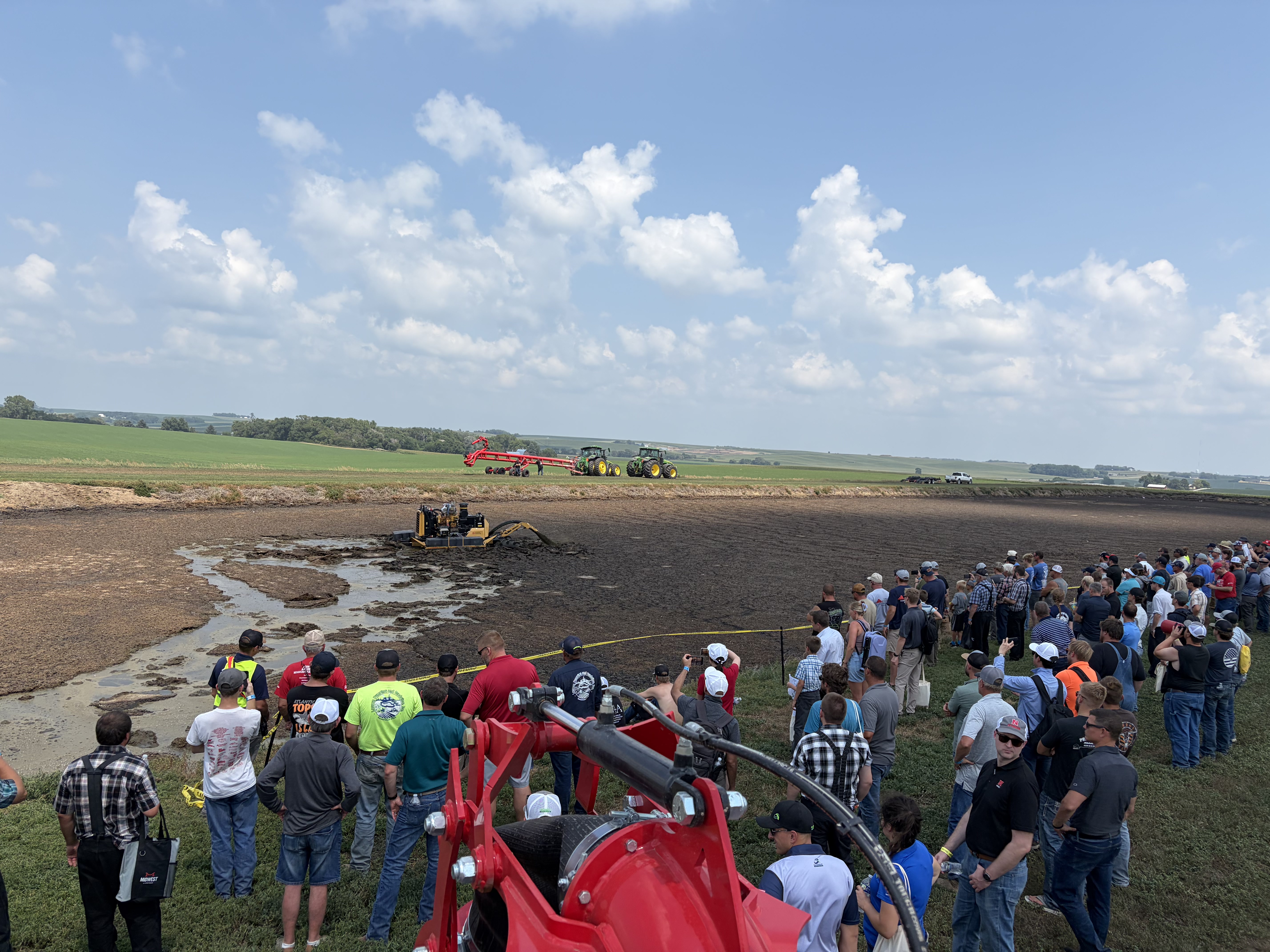 Crowd watches machinery demonstrating in a manure pit under a blue sky.