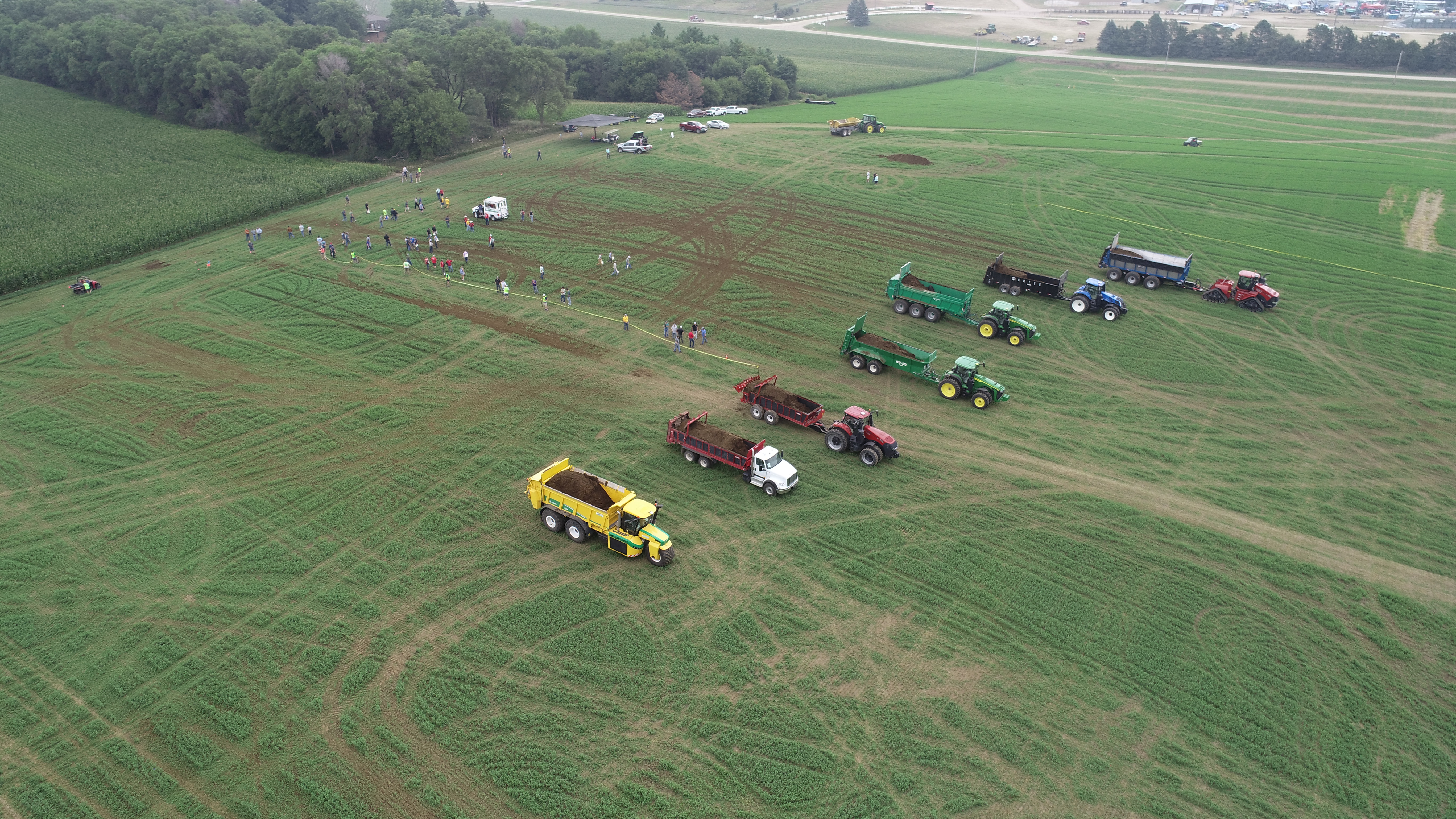 Solid manure spreaders lined up in an alfalfa field with scattered people and trees in the background.