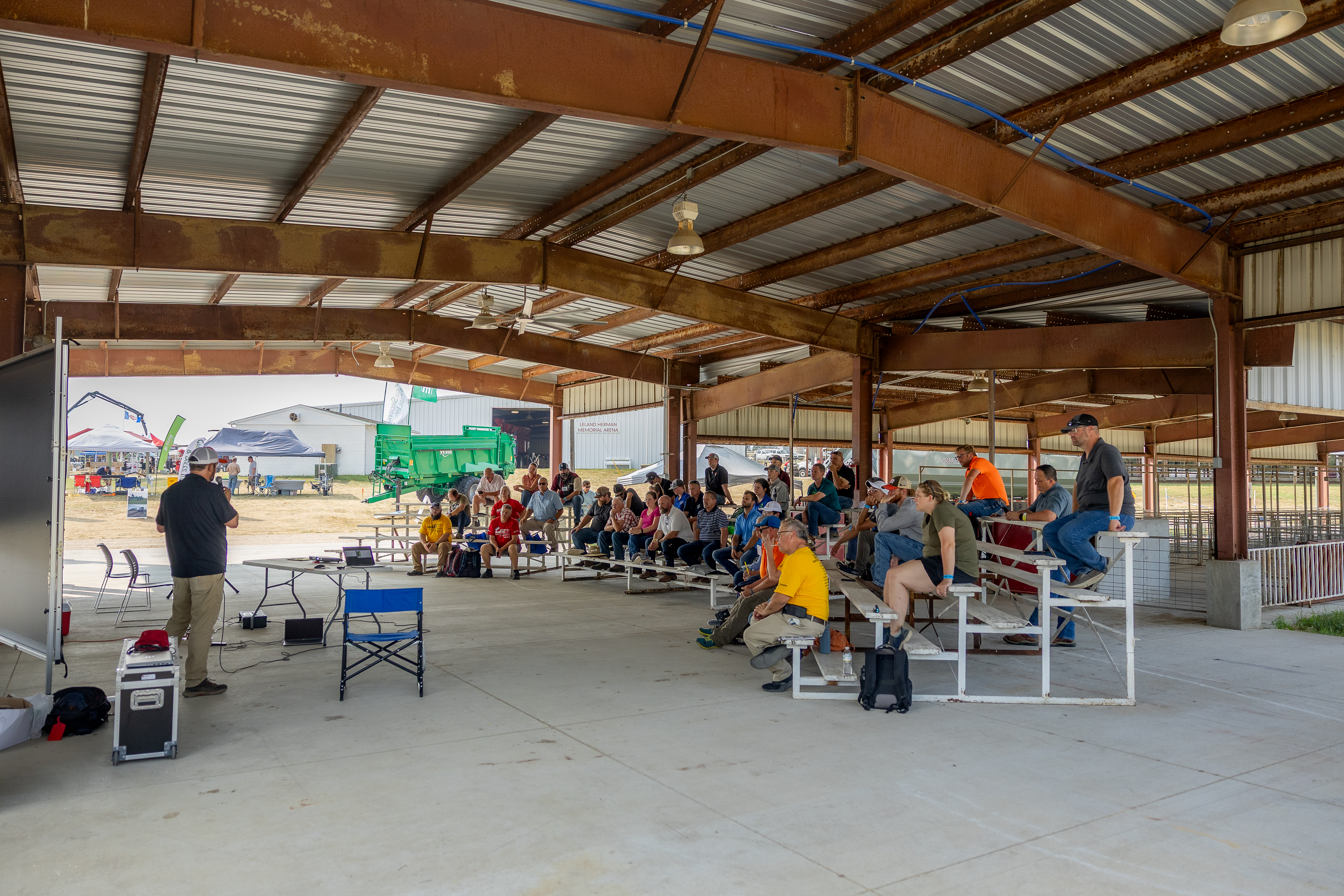 Audience seated under a metal pavilion, listening to a presentation. Machinery visible in the background.