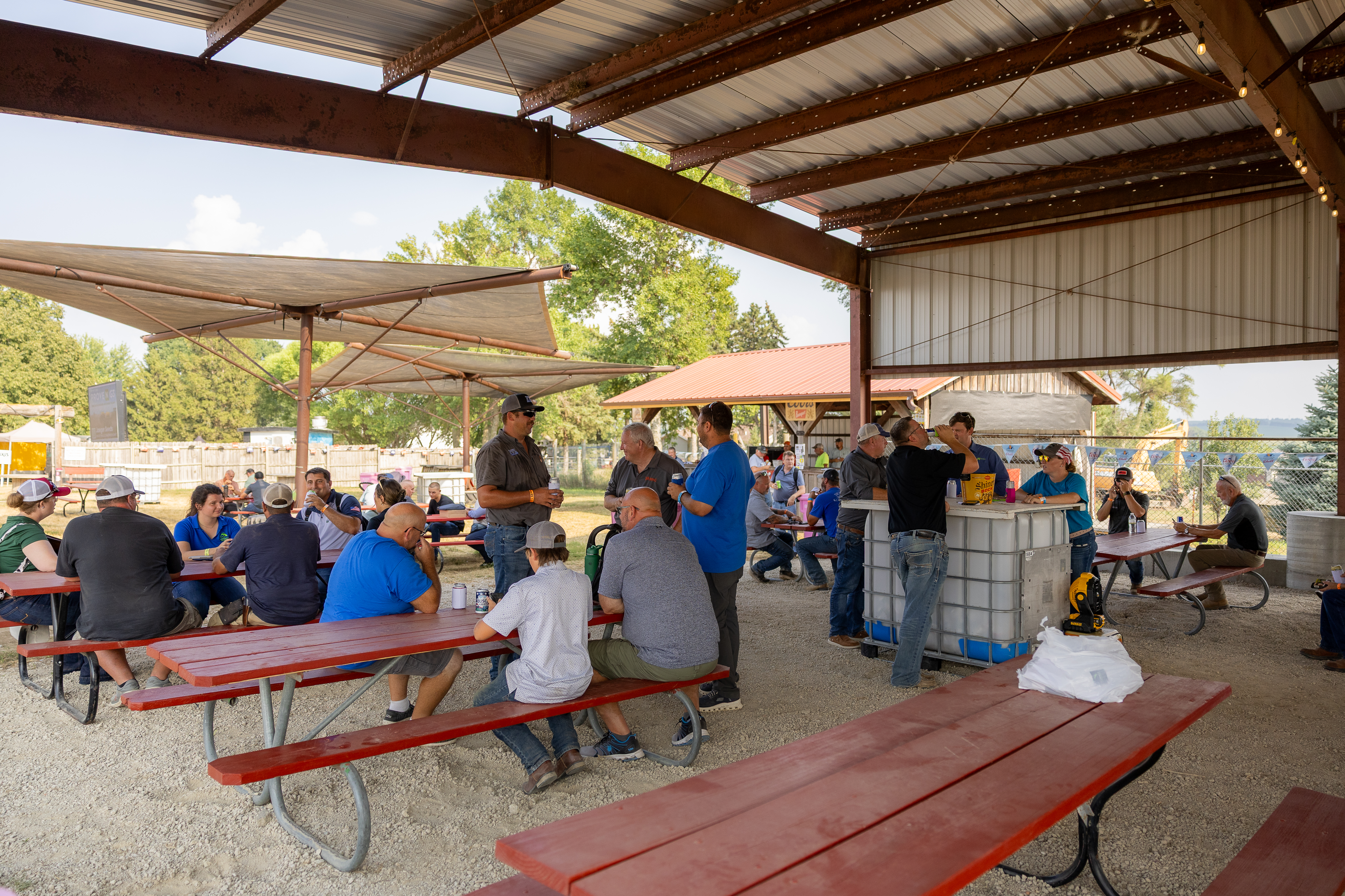 People gathered under a metal pavilion with picnic tables on a sunny day.