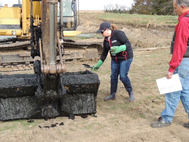 person sampling lagoon sludge from an excavator bucket