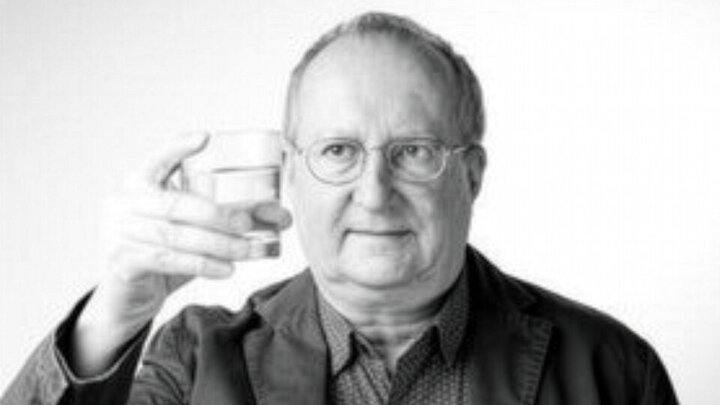 Man holding a glass of water, wearing a jacket and dotted shirt, black and white photo.